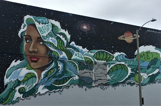 A vibrant mural featuring a woman's face with long, flowing hair that transforms into ocean waves, dotted with phrases like 'beauty of the wild' and 'be with me.' The backdrop is a starry night sky with celestial bodies, including a galaxy and a planet. A streetlamp stands in the foreground.