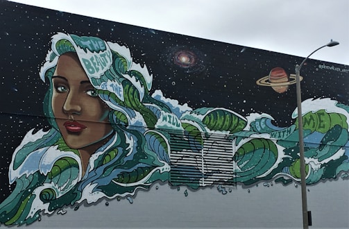 A vibrant mural featuring a woman's face with long, flowing hair that transforms into ocean waves, dotted with phrases like 'beauty of the wild' and 'be with me.' The backdrop is a starry night sky with celestial bodies, including a galaxy and a planet. A streetlamp stands in the foreground.