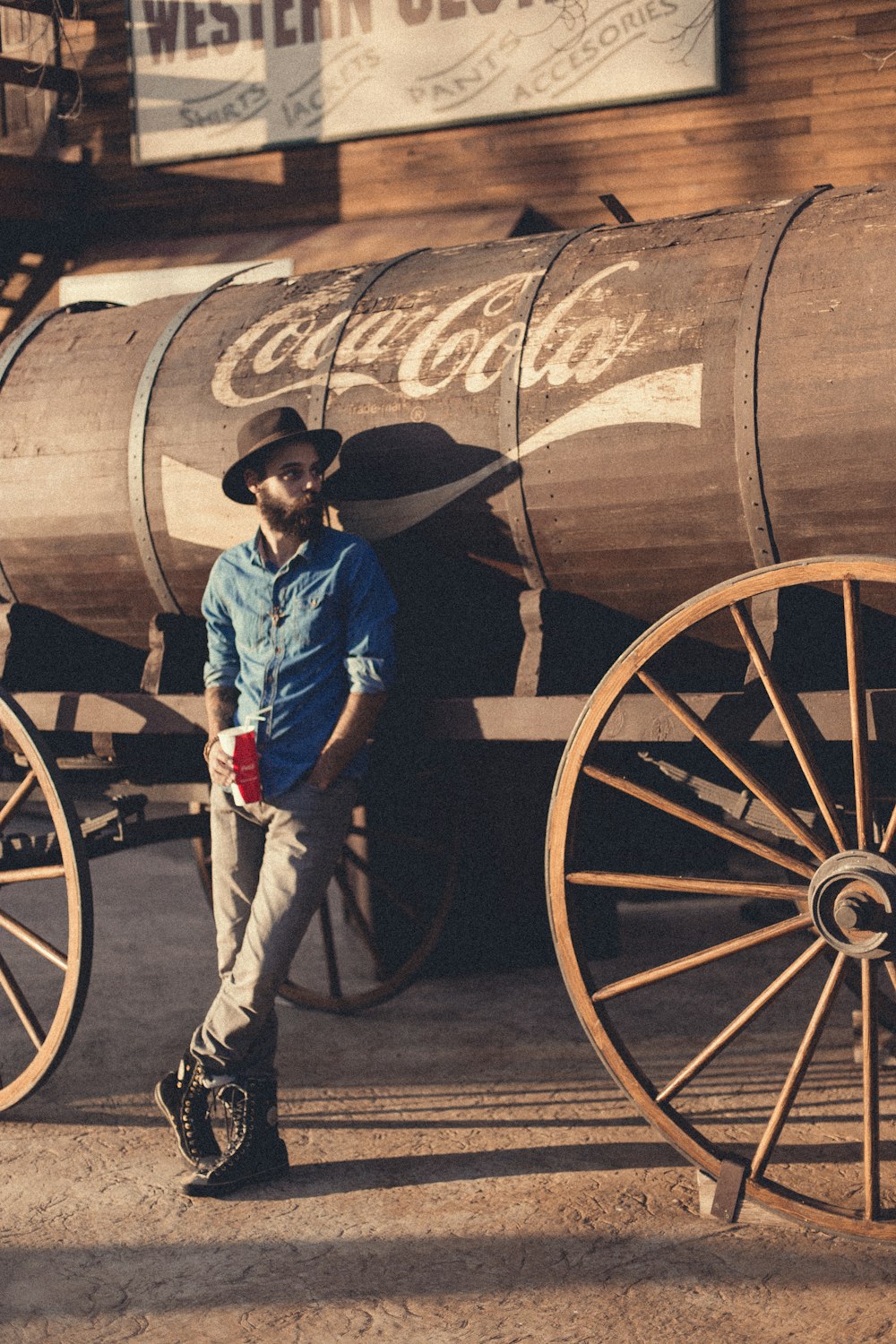 man in blue chambray sports shirt leaning on Coca-Cola barrel