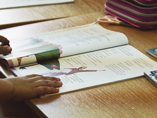 A child's hands are shown resting on an open book placed on a wooden table. The book contains text and images, including a photo of a person performing a yoga pose. Nearby, there is a pink-striped pencil case and a pair of scissors.