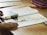 A child's hands are shown resting on an open book placed on a wooden table. The book contains text and images, including a photo of a person performing a yoga pose. Nearby, there is a pink-striped pencil case and a pair of scissors.