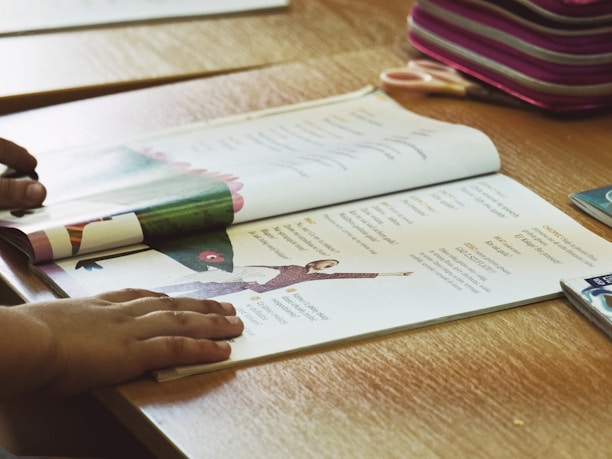 A child's hands are shown resting on an open book placed on a wooden table. The book contains text and images, including a photo of a person performing a yoga pose. Nearby, there is a pink-striped pencil case and a pair of scissors.