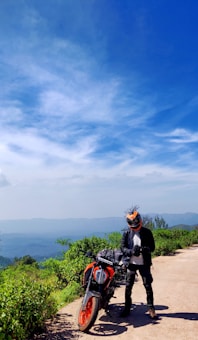 A person wearing a helmet and motorcycle gear stands beside a parked motorcycle on a scenic path with lush greenery. The sky above is bright blue, and distant hills are visible, creating a sense of open space and adventure.
