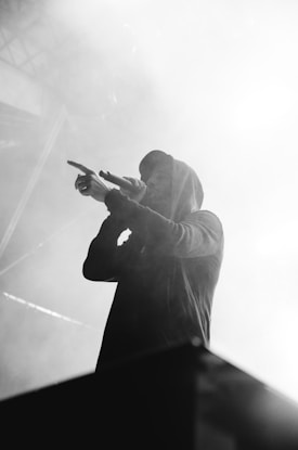 A person in a hoodie is performing on stage, holding a microphone and gesturing with their hand. The scene is enveloped in dramatic, heavy smoke and lighting, creating a moody atmosphere.