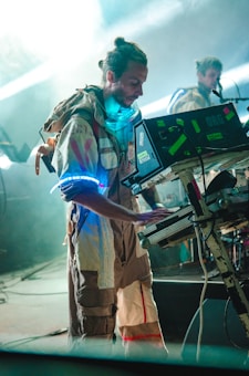 A musician dressed in an astronaut-like outfit is playing a keyboard or synthesizer on stage. The setting appears futuristic with glowing elements on the costume and equipment. The background is filled with smoke or a foggy ambiance, and another person in similar attire is seen in the background.