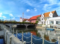 A scenic canal surrounded by historical buildings with red-tiled roofs and a clear blue sky above. The canal is lined with a decorative barrier, and there is a bridge connecting the two sides. There's a person visible near the canal, and palm trees add to the tropical feel.