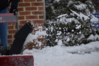 A snowblower is in action, throwing a flurry of snow into the air. The background features a brick wall and snow-covered greenery. A person is operating the machine, visible from the waist down.