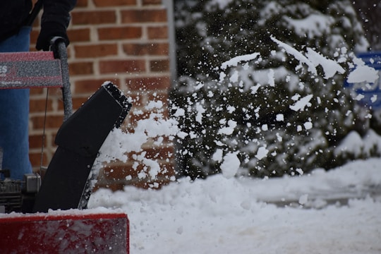 A snowblower is in action, throwing a flurry of snow into the air. The background features a brick wall and snow-covered greenery. A person is operating the machine, visible from the waist down.