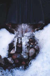 Icicles form from a dark, cylindrical drain pipe, surrounded by an accumulation of snow. Beneath, a bed of small, multicolored pebbles is partially visible, adding texture and contrast to the icy scene.