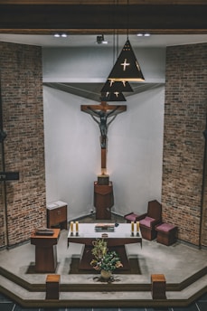 A church interior featuring a large crucifix mounted on the wall above an altar. The altar is adorned with several candles, a book, and a floral arrangement. The surrounding decor includes brick walls, wooden furniture, and a suspended light fixture with cross-shaped cutouts.