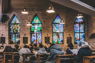 A diverse group of people worshiping together with warm sunlight streaming through stained glass windows.