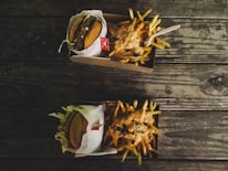 Two cardboard containers are placed on a rustic wooden table. One contains a hamburger wrapped in paper, while the other is filled with French fries topped with a creamy sauce. A plastic fork is resting on the fries.