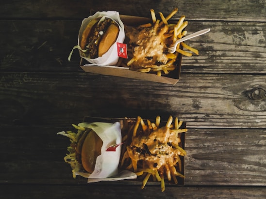 Two cardboard containers are placed on a rustic wooden table. One contains a hamburger wrapped in paper, while the other is filled with French fries topped with a creamy sauce. A plastic fork is resting on the fries.