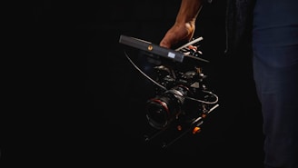 A person is holding a professional camera rig featuring a lens and attached handle, set against a dark background.
