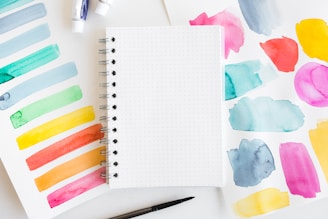 A spiral notebook with dotted pages is placed on a surface surrounded by sheets of paper displaying various watercolor swatches in different colors. Two small tubes of paint and a black paintbrush are also visible.