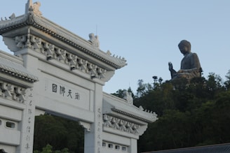 A large white traditional Chinese-style archway with intricate carvings stands in the foreground. Behind it, a massive bronze statue of a seated Buddha on a hill is visible, surrounded by trees under a clear blue sky.