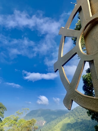 A large metal wheel sculpture is positioned against a backdrop of a clear blue sky, with a few scattered white clouds. In the distance, a lush, green mountain range and forest landscape adds depth and vibrancy to the scene.
