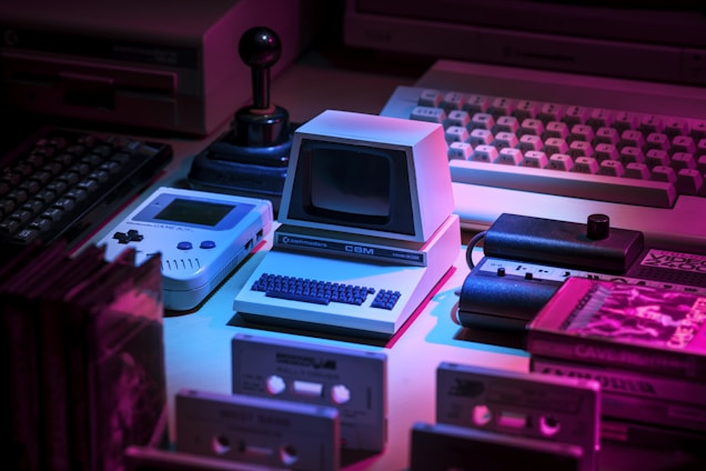A collection of vintage technology including a small Commodore computer, a Game Boy, an old joystick, and cassette tapes, all arranged under dim, colored lighting creating a nostalgic atmosphere. The overall setup gives a retro vibe with elements from 80s and 90s technology.