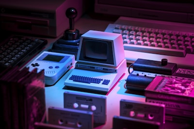 A collection of vintage technology including a small Commodore computer, a Game Boy, an old joystick, and cassette tapes, all arranged under dim, colored lighting creating a nostalgic atmosphere. The overall setup gives a retro vibe with elements from 80s and 90s technology.