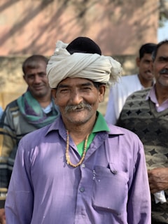 A group of men standing together, with one man in the foreground wearing a purple shirt and a turban. He has a mustache and a necklace with beads. The other men are dressed casually and are smiling in the background.
