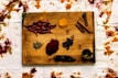 A wooden board displays a variety of spices, including whole dried red chilies, cinnamon sticks, a small wooden pestle and mortar, turmeric powder, and other loose spices. Surrounding the board are scattered piles of vibrant spices on a white fabric, adding an artistic and rustic feel to the composition.