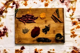 A wooden board displays a variety of spices, including whole dried red chilies, cinnamon sticks, a small wooden pestle and mortar, turmeric powder, and other loose spices. Surrounding the board are scattered piles of vibrant spices on a white fabric, adding an artistic and rustic feel to the composition.