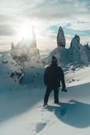 A person stands in a snowy landscape with jagged ice formations under a bright sun. The individual appears to be holding a camera and is dressed in dark winter clothing, with deep footprints trailing behind. The sky is partially cloudy with patches of blue.