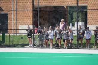 A group of women standing behind a chain-link fence, wearing sports uniforms with numbers visible. They are holding lacrosse sticks and standing on a field with a green surface. A brick building is visible in the background, and there's a person in colorful clothing standing out among them.