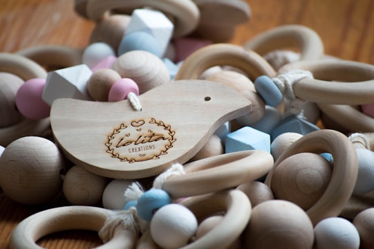 A wooden bird-shaped ornament with engraved text and decorative motifs is surrounded by an assortment of wooden rings and multicolored beads in soft pastel shades. The beads are geometric in shape, including spheres and hexagons, strung together with cords.