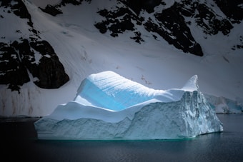 A massive iceberg with sharp, angular edges floats majestically in calm, dark waters surrounded by snow-covered cliffs. The sunlight casts a luminous blue glow on the iceberg's surface, creating a striking contrast against the dark background.