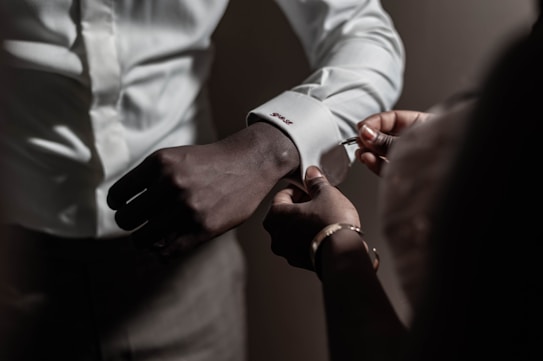 Two hands are assisting in fastening a cufflink on a white dress shirt. The lighting is subdued, focusing on the hands and the sleeve details.