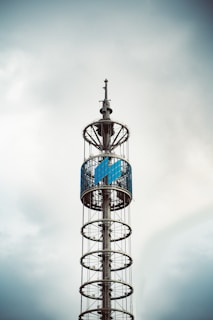 A tall, modern tower with circular platforms stacked vertically, featuring a large, enclosed section with blue panels near the top. The structure is silhouetted against a cloudy sky, giving it an industrial and sleek appearance.