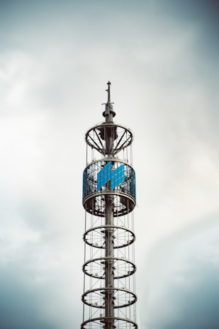 A tall, modern tower with circular platforms stacked vertically, featuring a large, enclosed section with blue panels near the top. The structure is silhouetted against a cloudy sky, giving it an industrial and sleek appearance.