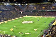 A large soccer stadium filled with a crowd of spectators, showcasing vibrant yellow and black team colors. The field is set for the match with multiple large flags being waved by individuals on the pitch. The stands are packed, creating a lively atmosphere as fans eagerly await the game.