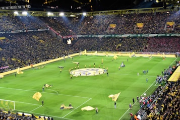 A large soccer stadium filled with a crowd of spectators, showcasing vibrant yellow and black team colors. The field is set for the match with multiple large flags being waved by individuals on the pitch. The stands are packed, creating a lively atmosphere as fans eagerly await the game.