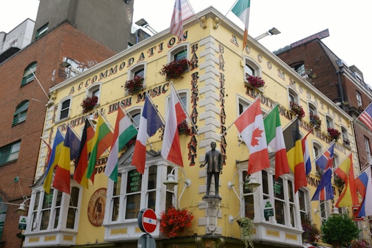 A vibrant corner building adorned with numerous international flags hanging from the facade. The structure features yellow walls with signs advertising a traditional Irish music bar and restaurant. Flower baskets are placed on the ledges, adding a touch of color. A small statue of a man is positioned above the entrance alongside classic architectural elements like windows with white shutters.
