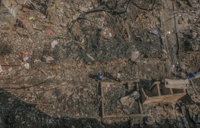 Aerial view of a large, cluttered area filled with debris and charred remnants. The ground appears burnt, with scattered pieces of wood, metal, and various discarded items. Two people can be seen walking through the debris, indicating activity. The area seems to have suffered a fire or some form of destruction, with remains of structures visible amidst the rubble.