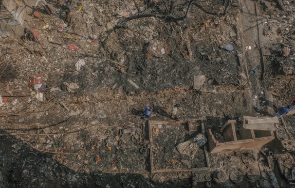 Aerial view of a large, cluttered area filled with debris and charred remnants. The ground appears burnt, with scattered pieces of wood, metal, and various discarded items. Two people can be seen walking through the debris, indicating activity. The area seems to have suffered a fire or some form of destruction, with remains of structures visible amidst the rubble.