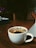 A white cup filled with herbal tea is placed on a wooden table. Mint leaves are visible floating in the tea. A laptop is partially visible to the right, and in the background, there is a soft focus on furniture, with pillows and dim, cozy lighting.