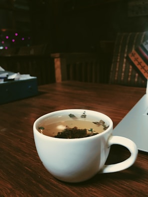 A white cup filled with herbal tea is placed on a wooden table. Mint leaves are visible floating in the tea. A laptop is partially visible to the right, and in the background, there is a soft focus on furniture, with pillows and dim, cozy lighting.