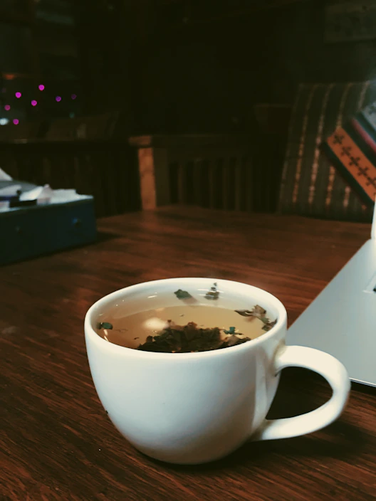 A steaming cup of yaupon tea resting on a wooden table with fresh herbs scattered around.