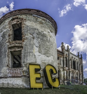 A vintage, partially ruined brick building with a cylindrical structure is set against a blue sky with scattered clouds. Large yellow letters with a dotted pattern spell 'EC' at the base of the structure. The building features arched windows and detailed stonework, conveying a sense of historical architecture.