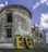A vintage, partially ruined brick building with a cylindrical structure is set against a blue sky with scattered clouds. Large yellow letters with a dotted pattern spell 'EC' at the base of the structure. The building features arched windows and detailed stonework, conveying a sense of historical architecture.