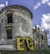 A vintage, partially ruined brick building with a cylindrical structure is set against a blue sky with scattered clouds. Large yellow letters with a dotted pattern spell 'EC' at the base of the structure. The building features arched windows and detailed stonework, conveying a sense of historical architecture.