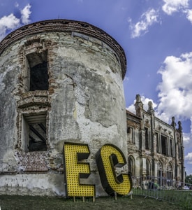 A vintage, partially ruined brick building with a cylindrical structure is set against a blue sky with scattered clouds. Large yellow letters with a dotted pattern spell 'EC' at the base of the structure. The building features arched windows and detailed stonework, conveying a sense of historical architecture.