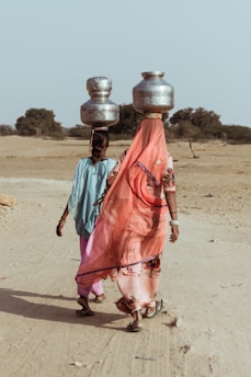 Two women walk along a dusty path in a barren landscape, carrying large metal pots on their heads. They wear traditional colorful clothing, with one woman in a bright orange and pink sari and the other in blue and pink attire. The background shows sparse vegetation and a clear sky, suggesting an arid environment.