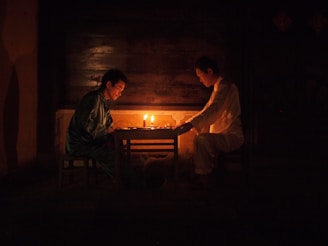 Two individuals are seated across from each other in a dimly lit room, with a warm glow from candles on a table between them. They appear to be engaged in a quiet, intimate conversation or activity. The wooden background suggests a rustic or traditional setting.