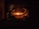 Two individuals are seated across from each other in a dimly lit room, with a warm glow from candles on a table between them. They appear to be engaged in a quiet, intimate conversation or activity. The wooden background suggests a rustic or traditional setting.