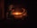 Two individuals are seated across from each other in a dimly lit room, with a warm glow from candles on a table between them. They appear to be engaged in a quiet, intimate conversation or activity. The wooden background suggests a rustic or traditional setting.