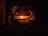 Two individuals are seated across from each other in a dimly lit room, with a warm glow from candles on a table between them. They appear to be engaged in a quiet, intimate conversation or activity. The wooden background suggests a rustic or traditional setting.